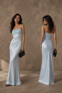 Two views of a woman modeling a VOGSION Alicia Light Blue Beaded-Fringe Petite Maxi Dress, standing against a textured warm brown studio backdrop. One view shows the front and the other shows the back, and each model holds a small black clutch.