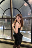 Woman in a Vogsion black corset draped mini dress standing in front of a large window with a view of a street.