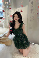 Model posing in a romantic cottagecore green dress in a flower-filled room, showing the flared mini skirt.