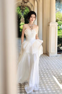 Vogsion Seraphina Royale luxury cream silk gown with peplum lace detailing and tiered skirt, worn by a woman in a Mediterranean villa hallway with classical arches.