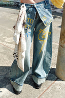 Model wearing VOGSION “Be Wild” graffiti wide-leg denim pants with yellow lettering, relaxed low-rise fit, and artistic street style — paired with black bralette and chunky knit cardigan for a bold Y2K look.