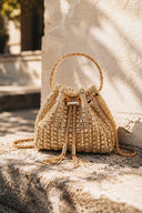 A woven straw bucket bag with a gold-tone metal handle and tassel details, resting on a textured stone ledge against a sunlit stucco wall.