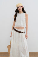 Woman wearing a white sleeveless top and long skirt with a yellow hat against a plain background