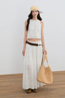 Woman wearing a white sleeveless top and long skirt with a beige bag, standing against a plain background.