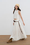 Woman wearing a white sleeveless top and long white skirt with brown boots against a white background