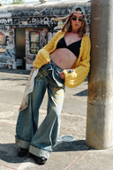 Model wearing VOGSION “Be Wild” graffiti wide-leg denim pants with yellow lettering, relaxed low-rise fit, and artistic street style — paired with black bralette and chunky knit cardigan for a bold Y2K look.