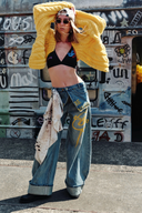 Model wearing VOGSION “Be Wild” graffiti wide-leg denim pants with yellow lettering, relaxed low-rise fit, and artistic street style — paired with black bralette and chunky knit cardigan for a bold Y2K look.