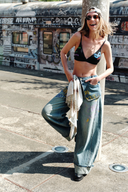 Model wearing VOGSION “Be Wild” graffiti wide-leg denim pants with yellow lettering, relaxed low-rise fit, and artistic street style — paired with black bralette and chunky knit cardigan for a bold Y2K look.