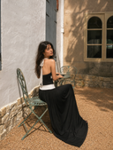 Rear angle of VOGSION Riviera Muse black halter backless maxi dress with plunging neckline, gold button waist detail and luxury Mediterranean courtyard setting.