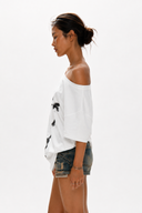 Petite model wearing the VOGSION ÉCLIPSE oversized white off shoulder graphic tee with abstract eye print. Breathable cotton loose fit summer t-shirt designed for petite women with a flattering off shoulder neckline that can also be worn as a boat neck, perfect for casual streetwear, travel, layering and everyday outfits.
