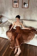 A woman sits on a white sofa in a room with wood-paneled walls and dark wood flooring, wearing a The Vogsion Liane Petite Wedding Guest Organza Ruffle Midi Dress with thin straps, a structured bodice, and a flowing sheer skirt with a decorative border.