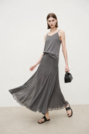 A woman modeling a VOGSION Celine High Waist Silk-Flow Maxi Skirt with a pleated skirt, paired with black sandals and holding a small black handbag, set against a minimalist off-white studio background.