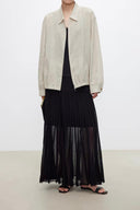 A model wearing a light beige oversized jacket over a black top and a VOGSION Celine High Waist Silk-Flow Maxi Skirt-black, paired with black slide sandals, set against a plain light gray studio background.