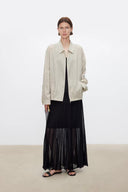 A model stands in a minimalist studio setting wearing a light beige oversized collared jacket over a black top and a VOGSION Celine High Waist Silk-Flow Maxi Skirt, paired with black sandals.