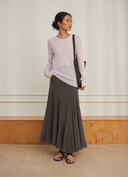 A woman stands in a minimalist indoor setting with a cream wall and light wood floor, wearing a light lavender long-sleeve top and a brown VOGSION Celine High Waist Silk-Flow Maxi Skirt , with a dark brown shoulder bag and black sandals.