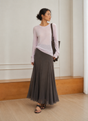 A woman stands in a minimalist indoor room wearing a light pink long-sleeve top and a VOGSION Celine High Waist Silk-Flow Maxi Skirt, with a dark shoulder bag and black sandals.