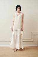 A woman stands in a minimalist room with cream walls and light wood flooring, wearing a cream ribbed sleeveless knit top and a matchi VOGSION Celine High Waist Silk-Flow Maxi Skirt, holding a small cream handbag.