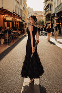 A woman wearing a VOGSION Noire Rosalie Glitter Rose Mermaid Maxi Dress stands on a sunlit city street, holding a dark umbrella. Pedestrians and storefronts are visible in the background.
