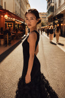 A woman wearing a VOGSION Noire Rosalie Glitter Rose Mermaid Maxi Dress stands on a sunlit city street, looking back over her shoulder. The background shows blurred pedestrians and storefronts in a real outdoor environment.