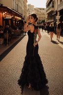 A woman stands on a sunlit city street wearing a VOGSION Noire Rosalie Glitter Rose Mermaid Maxi Dress and holding a single red rose. Café seating lines the left side of the street, with other pedestrians in the background.