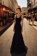 A woman stands on a sunlit city street wearing a VOGSION Noire Rosalie Glitter Rose Mermaid Maxi Dress with a fitted bodice and a voluminous textured skirt. Café awnings and other pedestrians are visible in the background.