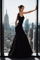A woman wearing VOGSION Noire Rosalie Glitter Rose Mermaid Maxi Dress with a textured, ruffled skirt stands by a large window overlooking a city skyline, with the Empire State Building visible in the background.