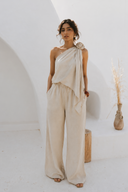 Model wearing the VOGSION Élan Quiet Luxury Linen Set featuring a premium linen one-shoulder top with handcrafted floral rosette detail and flowing wide-leg pants, styled for resort vacations, summer elegance, and effortless quiet luxury.