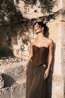 Vogsion “Aphrodite” Earth-Tone Draped Corset & Hip-Cutout Maxi Dress featuring a structured brown corset bodice with lace-up detailing, flowing earth-tone draped maxi skirt and subtle hip cutouts. Fashion model posing against sunlit Mediterranean stone walls beneath olive branches, captured in warm golden hour light with cinematic editorial styling, perfect for luxury resort wear, vacation outfits, destination weddings and elegant evening occasions.