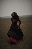 A woman in a black and red Victorian-style gothic dress stands in a dry grass field, her face obscured by a blindfold, with a hazy, moody sky in the background.