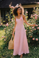 Woman wearing the Vogsion Last Sunset Rosette Linen Slip Dress in blush pink linen with a handcrafted rosette detail, full-length garden portrait surrounded by blooming roses, breathable summer linen maxi dress, wedding guest outfit, vacation dress, romantic cottagecore fashion, lightweight resort wear.