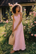 Full-length view of the Vogsion Last Sunset Rosette Linen Slip Dress featuring a blush pink linen slip silhouette and handcrafted rosette detail, romantic floral garden backdrop, cottagecore summer dress, lightweight linen maxi dress, elegant vacation outfit, bridal shower and wedding guest fashion.