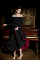 A woman modeling Vogsion Midnight Rose Off-Shoulder Mermaid Maxi Dress. She is standing barefoot in a dimly lit room with a wooden floor and a grand piano behind her, with a classical painting hanging on the wall.