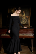 A woman wearing Vogsion Midnight Rose Off-Shoulder Mermaid Maxi Dress, a black off-the-shoulder long-sleeve mermaid gown stands beside a grand piano in a dark room, with a large framed painting behind her.