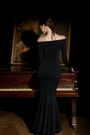 A woman wearing Vogsion Midnight Rose Off-Shoulder Mermaid Maxi Dress, a black off-the-shoulder long-sleeve mermaid gown stands with her back to the camera beside a dark wooden grand piano in a dimly lit room, with a large framed painting in the background.