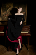 A woman wearing Vogsion Midnight Rose Off-Shoulder Mermaid Maxi Dress, a long black off-the-shoulder gown with a dramatic magenta-pink underlayer or train, seated on a wooden piano in a dark room with a large framed painting behind her.