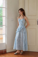 Full-length view of the Vogsion Sky Mist Blue Ruffle Maxi Dress featuring delicate blue watercolor floral print, asymmetric cascading ruffles, adjustable fabric straps, matching lining, and flowing chiffon silhouette for wedding guest and summer occasions.