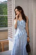 Sweet Ukrainian model wearing the Vogsion Sky Mist Blue Ruffle Maxi Dress beside a sunlit window. Soft blue floral print, cascading ruffle details, fully lined skirt, petite-friendly silhouette, perfect for summer weddings, garden parties, and elegant vacation styling.