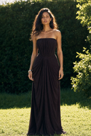 A woman stands outdoors on grass in front of a green hedge, wearing a Vogsion Strapless Ruched Maxi Dress - Dark Plum/Chocolate Wedding Guest Evening Gown with a gathered bodice and flowing skirt.