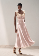 A model wearing Vogsion Sunset Aurelia Lace-Trimmed Liquid Satin Gown dress with ruffled lace detailing on the bodice and a flared skirt, paired with gold sandals, set against a neutral studio background.