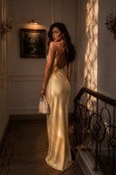 A woman stands on an indoor staircase landing wearing a Vogsion x Last Sunset Butter Yellow Satin Backless Slip Maxi Dress. She holds a small cream handbag in a warmly lit room with wall sconces, framed wall art, and a dark metal railing.