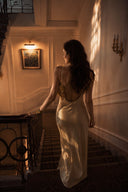 A woman in a Vogsion x Last Sunset Butter Yellow Satin Backless Slip Maxi Dress with a deep back walks down a staircase in a warmly lit, elegant interior.