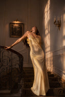 A woman in a Vogsion x Last Sunset Butter Yellow Satin Backless Slip Maxi Dress stands on an indoor staircase, leaning on a dark metal railing with her head tilted back in warm, sunlit light.