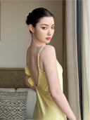 Woman in a Vogsion x Last Sunset Butter Yellow Satin Backless Slip Maxi Dress with Rosette Detail standing in a room with neutral decor