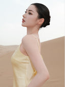 Woman in a Vogsion x Last Sunset Butter Yellow Satin Backless Slip Maxi Dress with Rosette Detail against a beige background