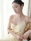 Woman wearing a Vogsion x Last Sunset Butter Yellow Satin Backless Slip Maxi Dress with Rosette Detail with floral details in a softly lit room.