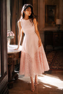 Woman wearing the VOGSION Rosalie Blush Lace Hem Midi Dress, elegant sleeveless pink midi wedding guest dress with laser cut lace hem in a luxury vintage hotel.