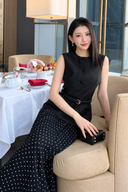 Close seated view of the black sleeveless dress showing its round neckline, covered-button detailing and included waist belt.