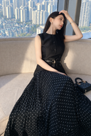 Side seated view of the regular-fit black polka-dot pleated dress, with its attached vest-style bodice and included belt.