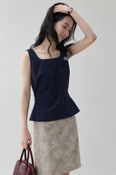 Odeline navy sleeveless rosette top with a flared hem over a taupe textured skirt, shown in a raised-arm pose.