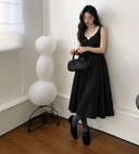 Black sleeveless cotton knit midi dress with ivory rosette neckline and gathered skirt, full-length model view beside a paper floor lamp.